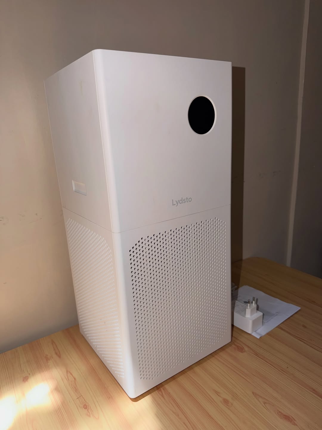 Air purifier putih berbentuk menara dengan panel lubang udara halus di bahagian bawah, berdiri atas meja kayu