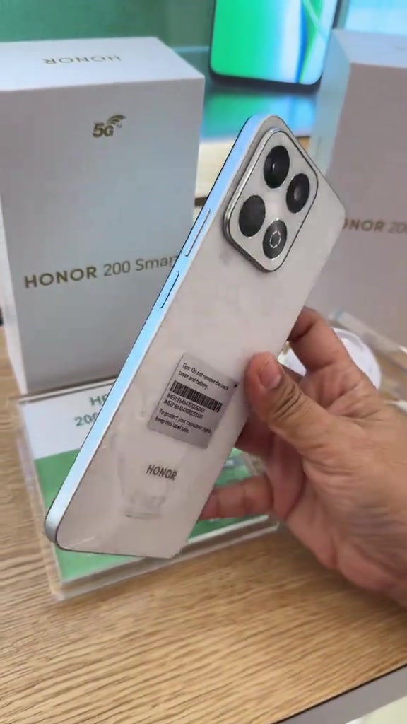 Unit phone HONOR warna putih dengan pelekat barcode IMEI di belakang dipegang tangan, dengan kotak HONOR di latar belakang