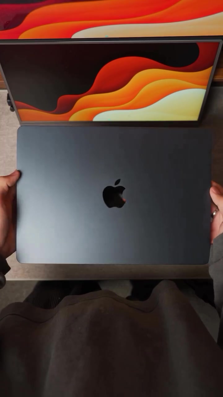 Penutup MacBook berwarna kelabu gelap dengan logo Apple dipegang dua tangan di atas meja