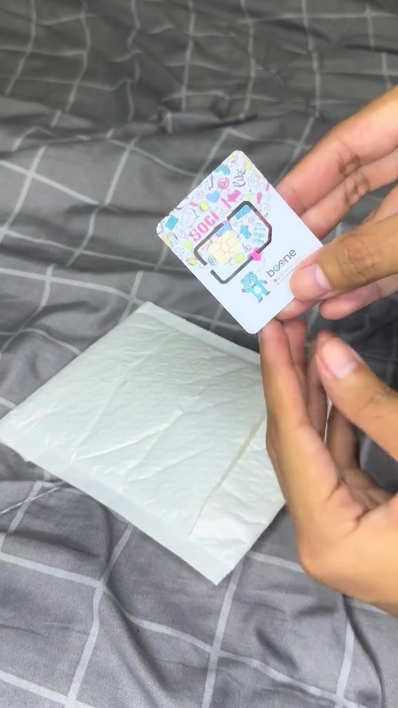 Tangan memegang kad SIM BeONE berwarna putih dengan rekaan berwarna-warni yang baru dikeluarkan daripada bungkusan bubble wrap di atas katil.