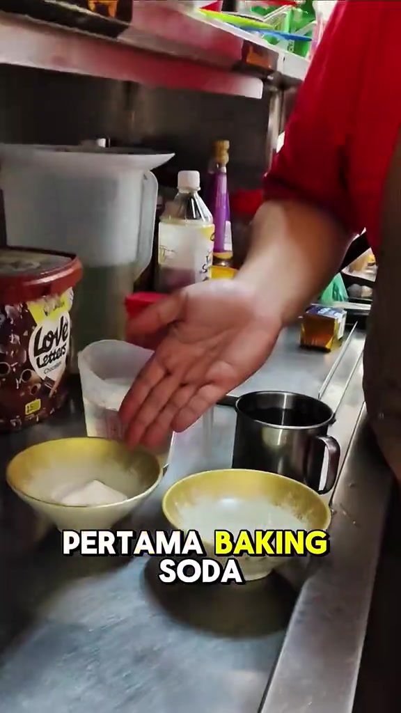 Mangkun berisi serbuk baking soda di atas meja dapur stainless bersama jag susu berisi air dan cawan keluli