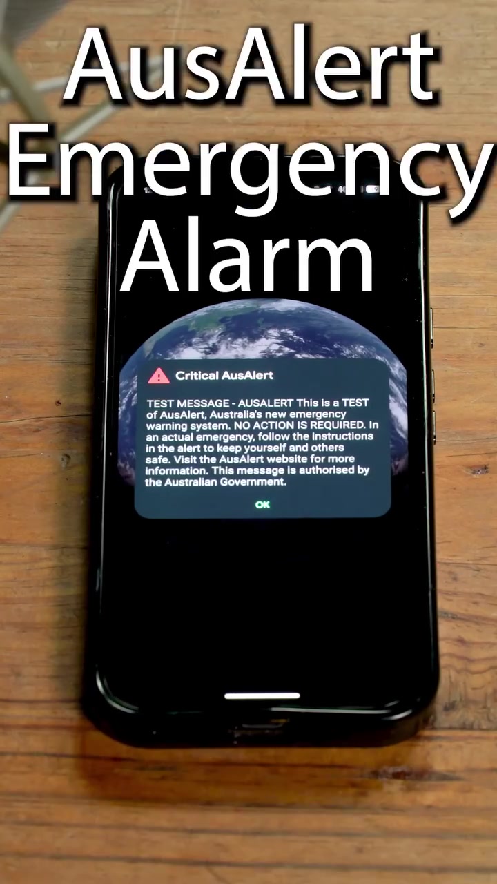 iPhone di atas meja kayu memaparkan pop-up Critical AusAlert dengan mesej ujian dan catatan NO ACTION IS REQUIRED