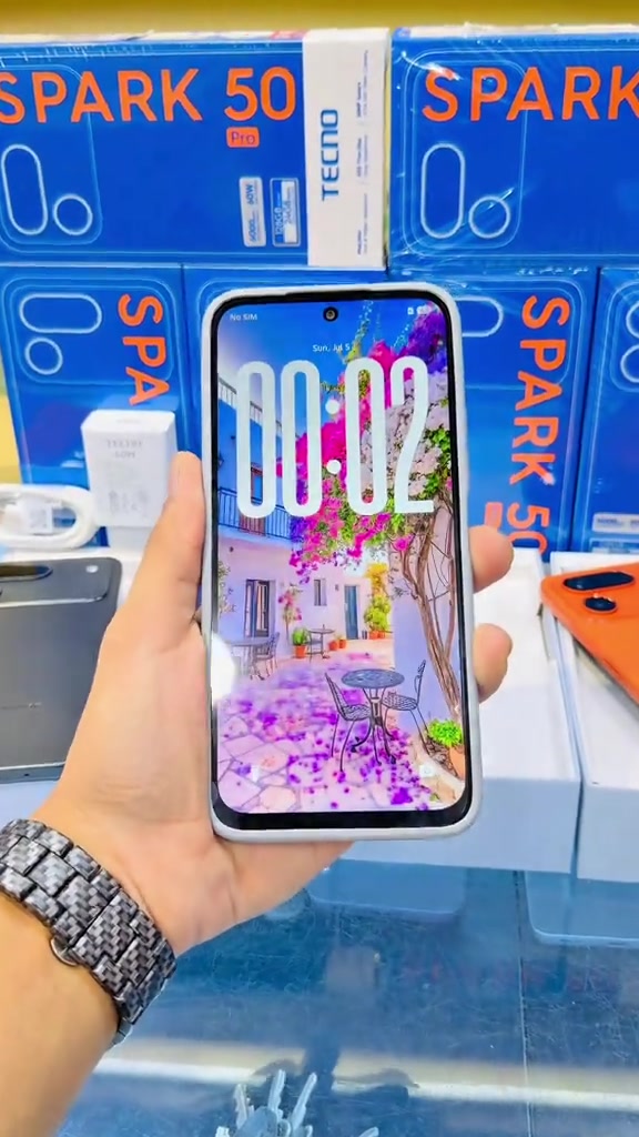 Tangan memegang Tecno Spark 50 Pro dengan skrin hidup di hadapan timbunan kotak runcit biru bertulis SPARK 50 Pro, bersama aksesori pengecas putih