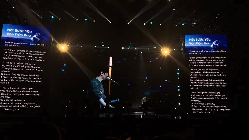 Sân khấu nhìn xa với đàn grand piano ở giữa, hai màn hình lời ca hai bên và bóng nghệ sĩ ngồi hát trong ánh đèn mờ