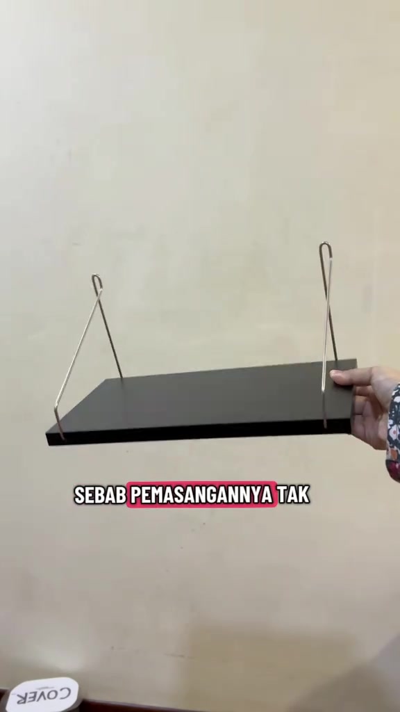 Papan rak hitam dengan bracket wayar besi nipis dipegang tangan di hadapan dinding cream