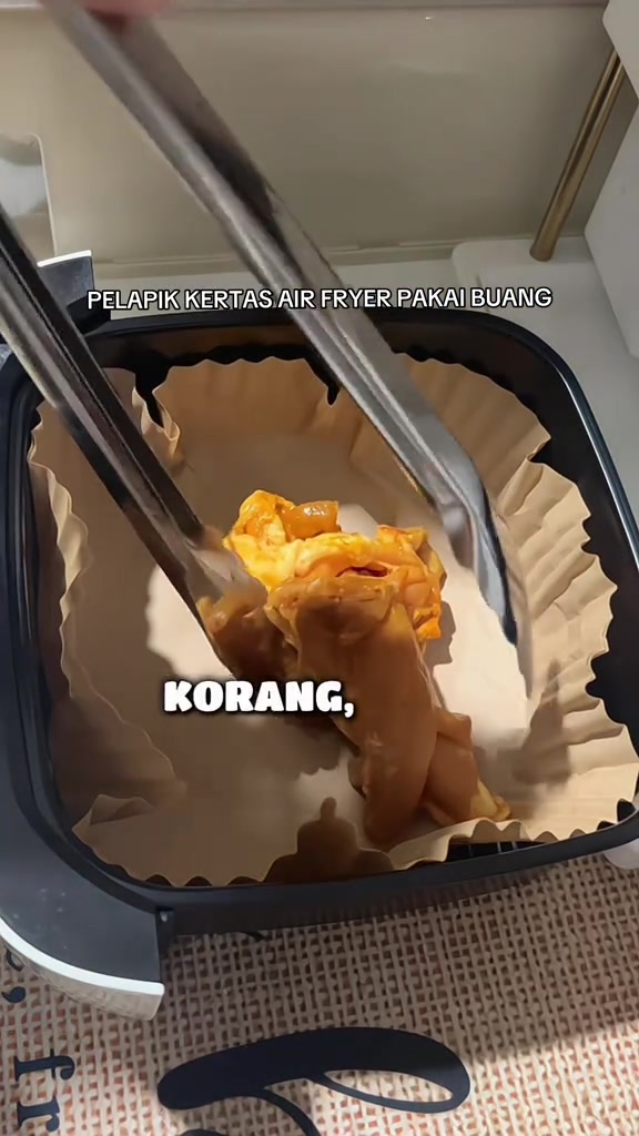 Penyepit logam meletakkan ayam berperap ke dalam bakul air fryer segi empat yang dilapik kertas parchment berlipat