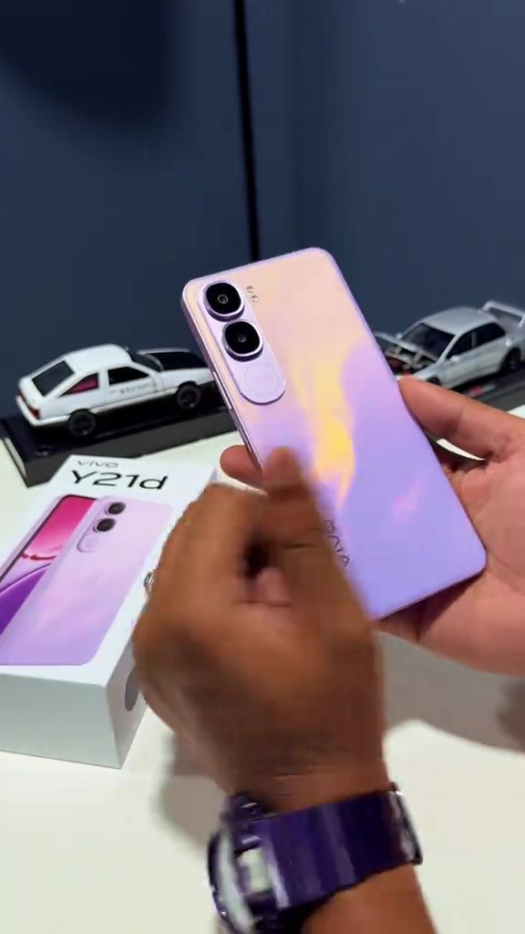 Kotak retail vivo Y21d dipegang hadapan kamera dengan gambar telefon ungu pada muka kotak, rak kedai di latar