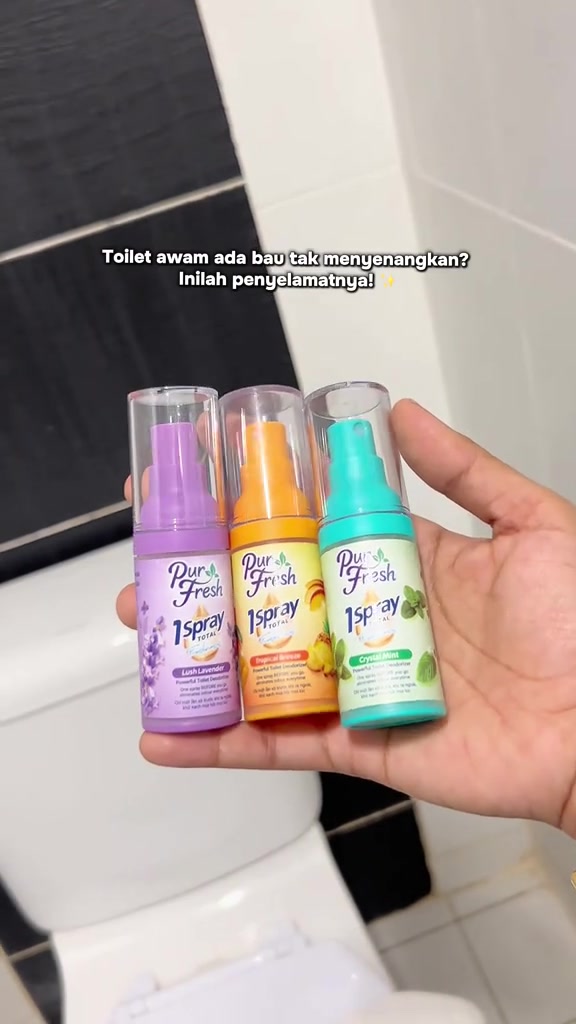 Tiga botol spray pewangi tandas Pur Fresh warna ungu, oren dan hijau dipegang di atas mangkuk tandas
