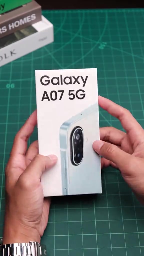 Kotak putih Samsung Galaxy A07 5G dipegang dua tangan di atas meja potong hijau