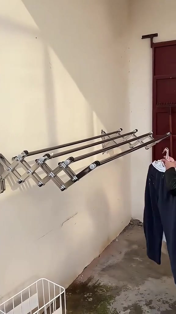 Rak jemuran lipat dinding stainless jenis accordion terbuka dengan empat bar, sebelah tangan menyangkut seluar gelap pada hujungnya