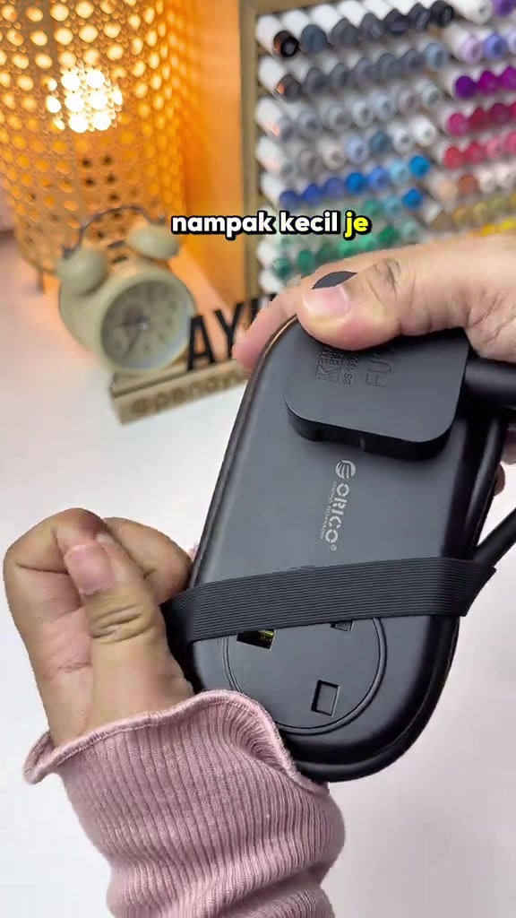 Power strip travel ORICO hitam dengan tali getah mengelilingi plug dan wayar yang dililit, dipegang dua tangan di atas meja