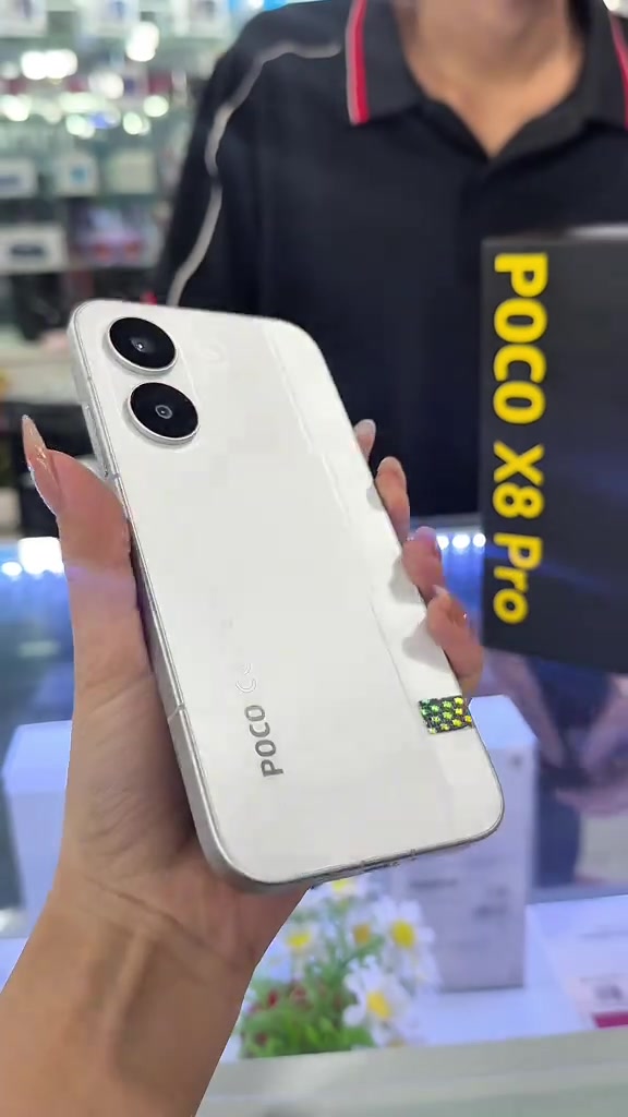 Mặt lưng trắng POCO X8 Pro với hai ống kính xếp dọc và logo POCO, phía sau là hộp máy màu đen chữ vàng