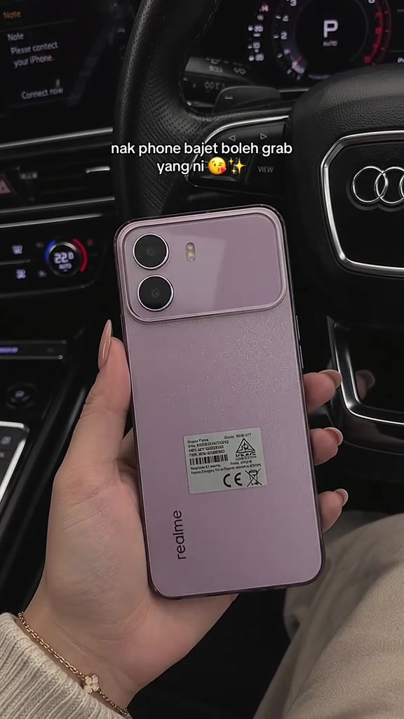 Telefon realme merah jambu dipegang dengan label model dan tanda CE masih melekat di belakang, latar dalam kereta