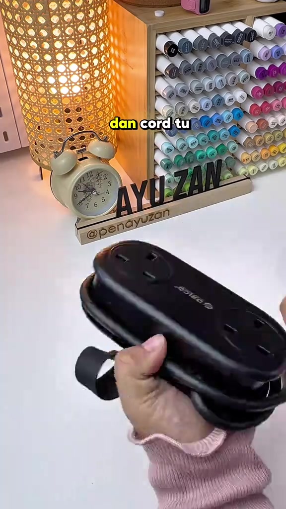 Power strip travel ORICO hitam dengan wayar dililit kemas di bawah badan unit, dua soket UK kelihatan pada muka atas, dipegang satu tangan