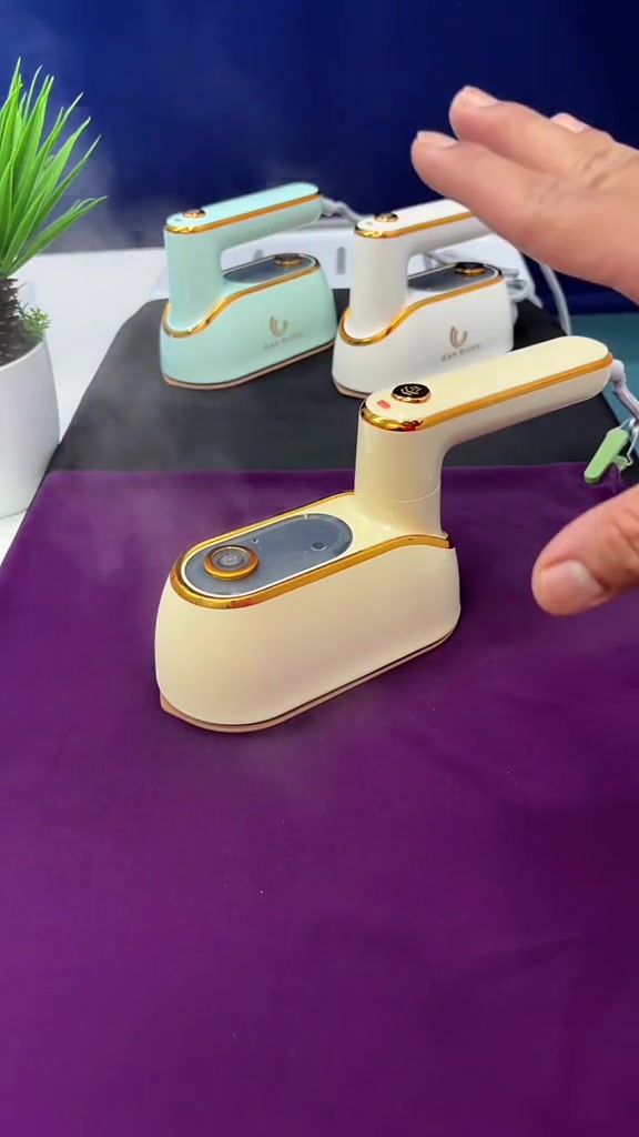 Tiga unit travel iron stim mini warna krim, putih dan hijau muda mengeluarkan wap di atas kain ungu