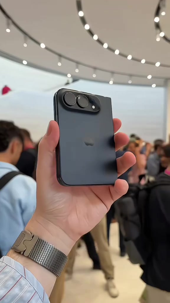 iPhone Duo màu xanh đen gập lại cầm trên một bàn tay giữa cửa hàng đông người, thấy rõ cụm hai camera sau nằm ngang