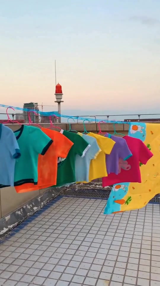 Barisan T-shirt berwarna-warni dan kain selimut kuning bergantung pada tali jemuran biru berlubang di rooftop terbuka