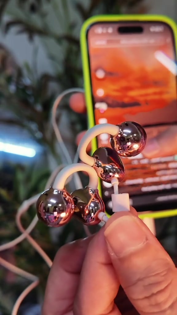 Earphone berwayar jenis klip telinga warna rose gold dipegang dekat kamera, dengan telefon di latar belakang