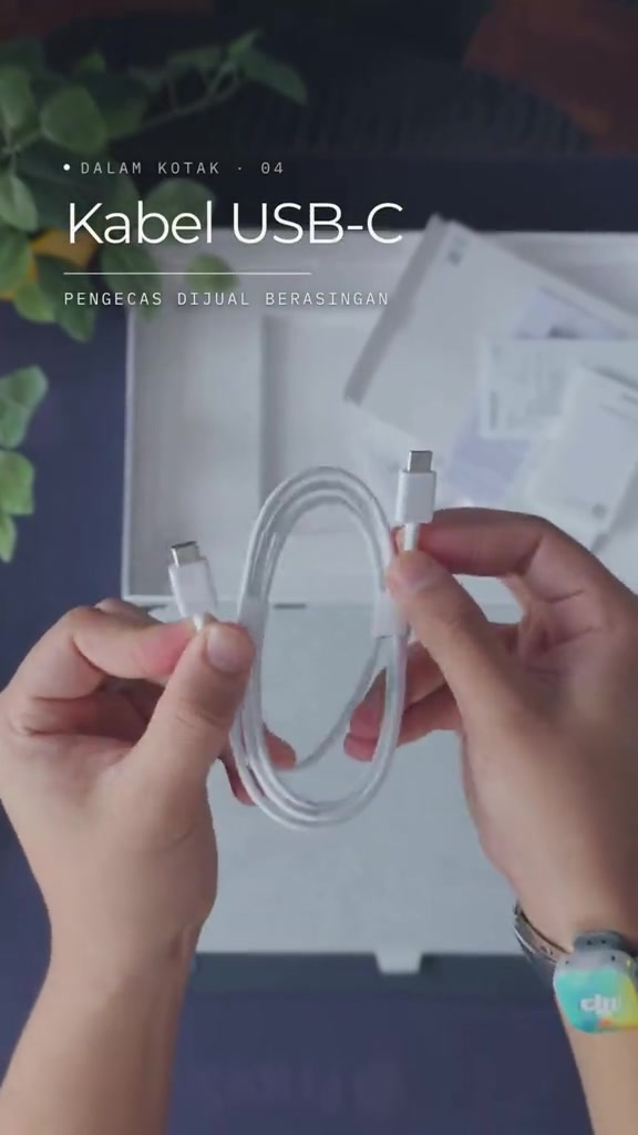 Kabel USB-C putih dipegang dua tangan di atas kotak tablet, dengan teks pada bingkai menyatakan pengecas dijual berasingan