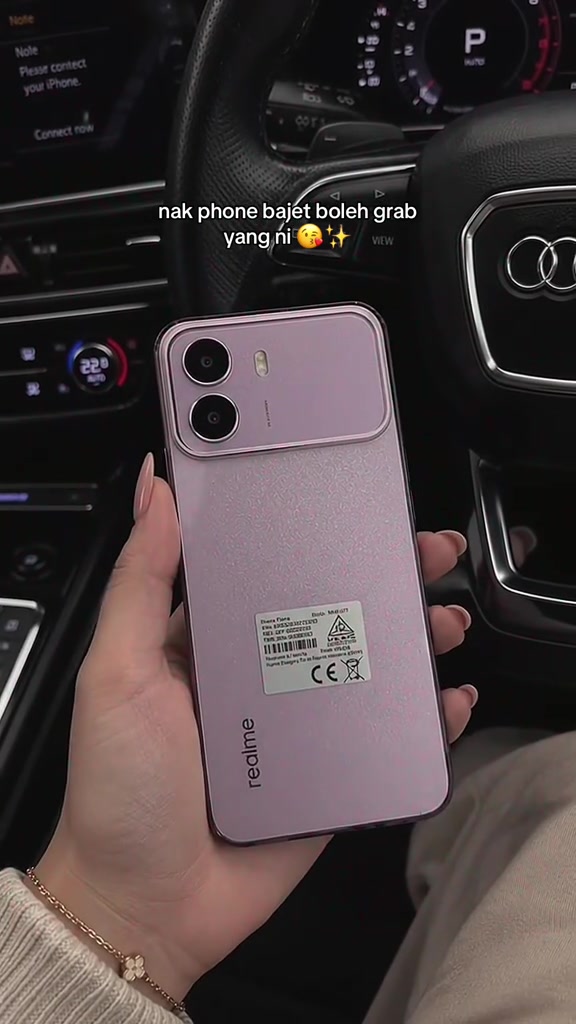 Bahagian belakang telefon realme berwarna merah jambu dipegang dalam kereta, dengan label model dan tanda peraturan masih melekat