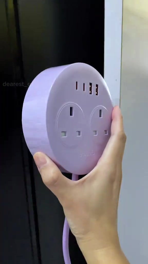 Power strip bulat ORICO warna lilac dipegang tangan — dua soket UK dan empat port USB pada muka atas, kabel lilac menjuntai ke bawah