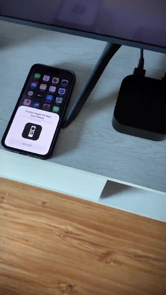 Unit Apple TV 4K di belakang TV dengan iPhone di sebelahnya memaparkan arahan kawalan Apple TV