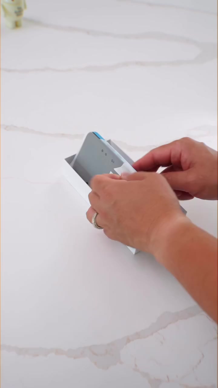 Tangan sedang mengeluarkan unit Nothing Phone (4b) dari kotak putihnya di atas meja putih