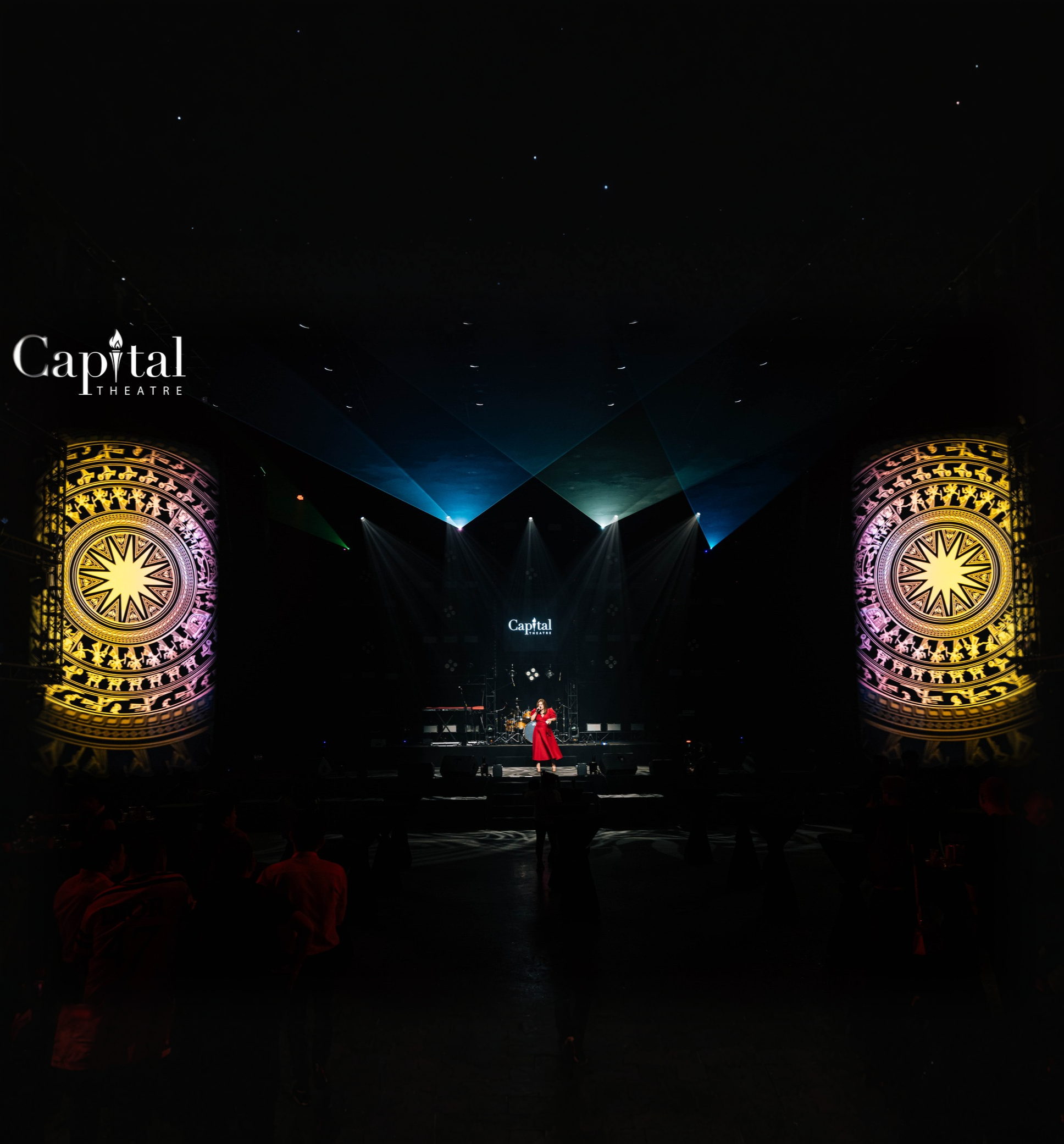 Không gian nhà hát Capital Theatre với hai cột màn hình LED họa tiết trống đồng rực sáng hai bên, sân khấu xa nhuộm ánh đèn xanh và bóng khán giả mờ phía trước
