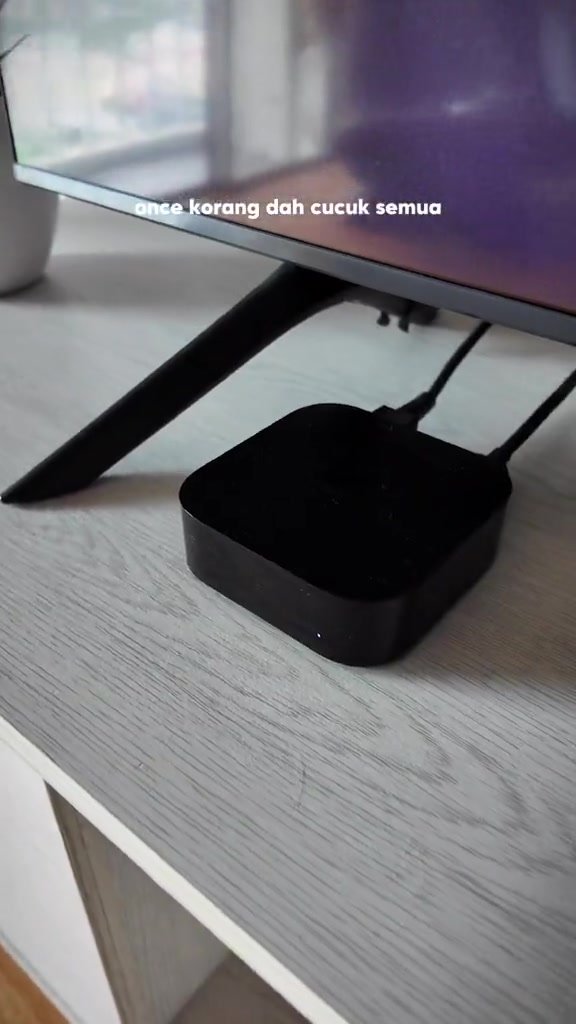 Unit Apple TV berwarna hitam disambung kabel di belakang TV di atas kabinet putih