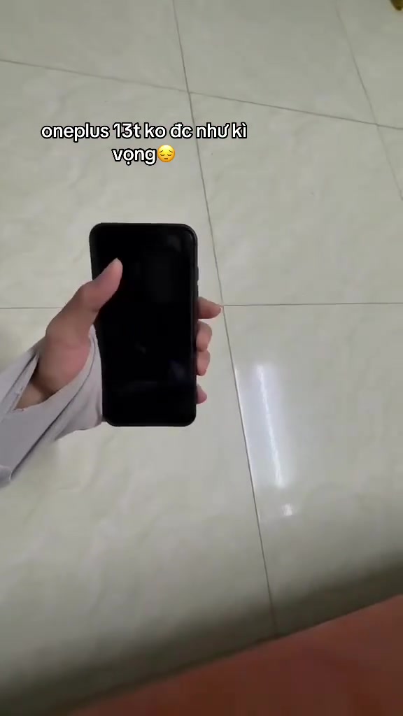 Bàn tay cầm OnePlus 13T màu đen màn hình tắt trên nền gạch men sáng