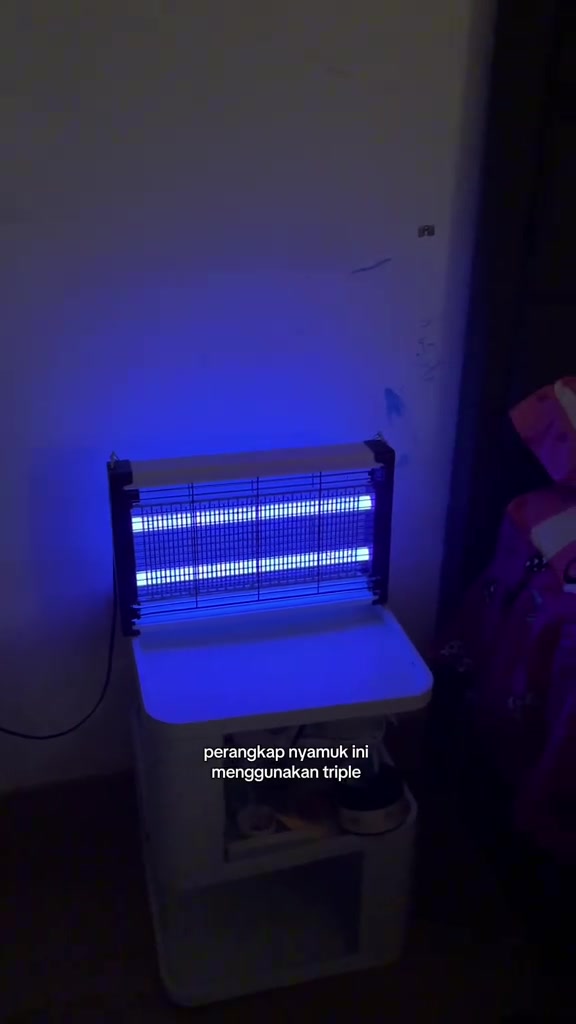 Perangkap nyamuk lampu UV bergrid menyala biru di atas meja sisi katil dalam bilik gelap