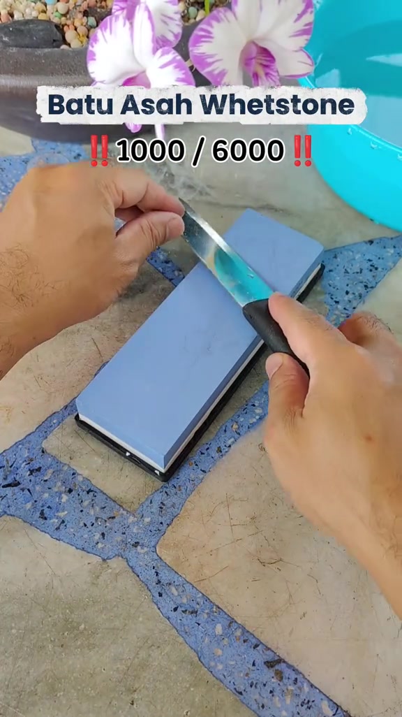 Dua tangan mengasah pisau kecil di atas batu asah dua muka berwarna biru dan putih bertanda 1000/6000