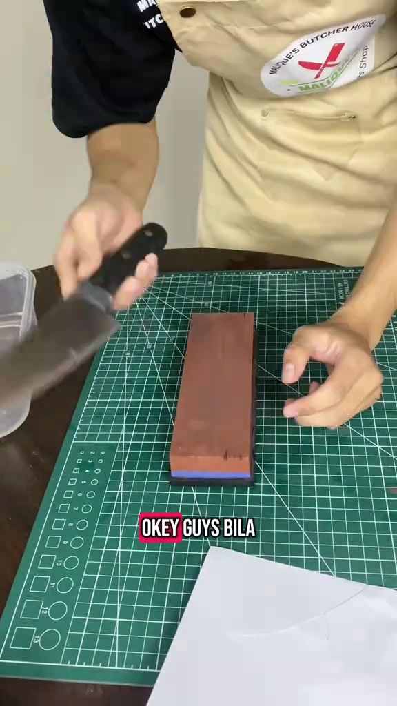 Tangan memegang pisau dapur di atas batu asah di atas tikar potong hijau dengan bekas air di sebelah