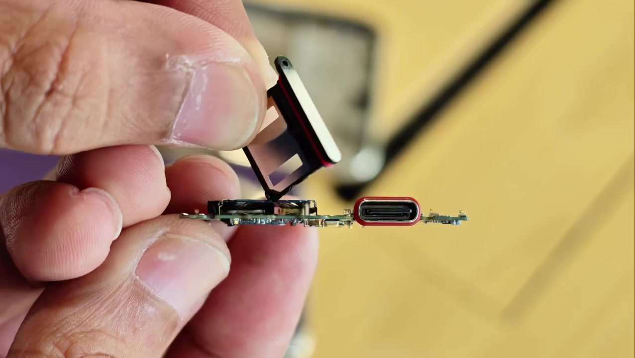 Jari memegang tray slot SIM dan papan litar telefon dengan gelang getah seal merah kelihatan mengelilingi port USB-C