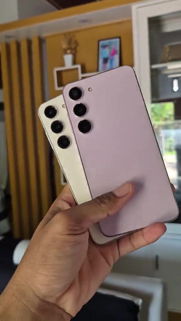 Dua unit Samsung Galaxy S23 warna lavender dan krim diperiksa dari sudut belakang di dalam tangan