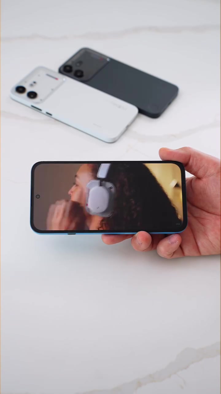 Dua unit Nothing Phone (4b) warna putih dan hitam diletak di atas meja, sementara satu unit biru di tangan memaparkan video pada skrin