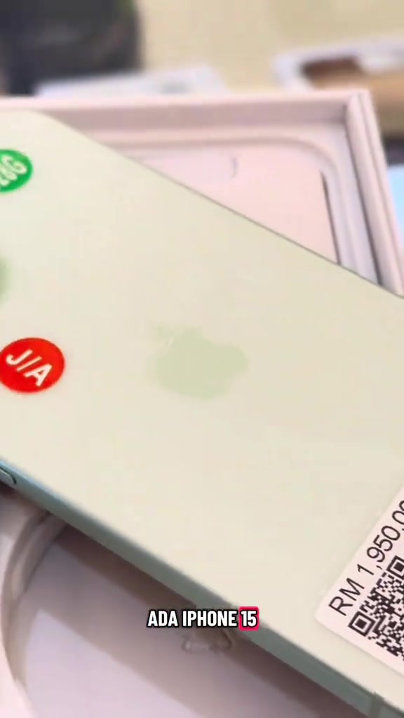 Bahagian belakang iPhone 15 warna hijau muda dengan pelekat bulat merah bertulis J/A dan tag harga bercetak RM1,950.00