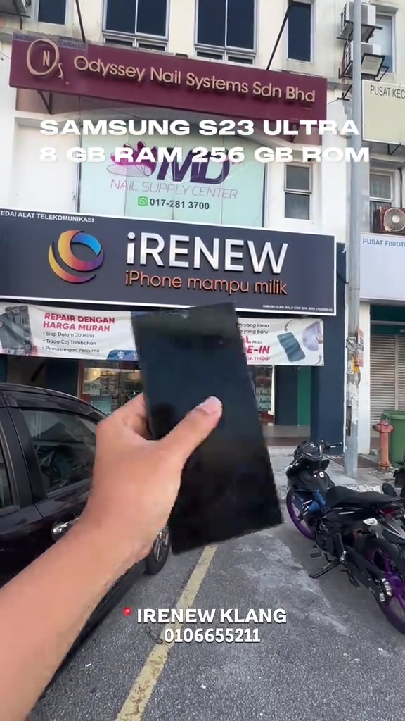 Depan kedai telefon terpakai iRenew di Klang dengan papan tanda besar, sebelah tangan memegang phone hitam di hadapan kedai
