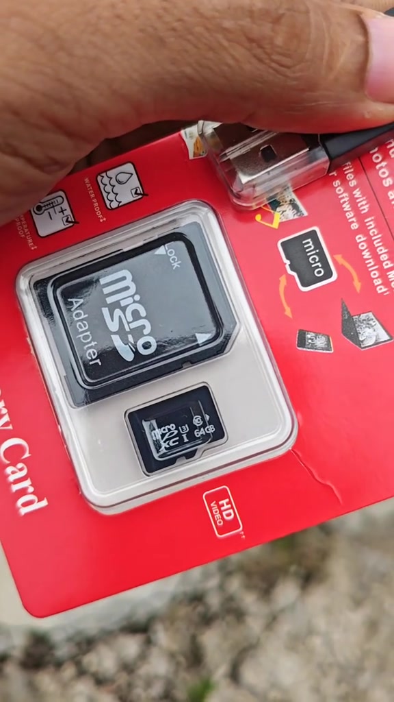 Tangan memegang pek blister merah tanpa jenama; kad microSD di dalamnya berlabel 64GB sebelah adapter SD