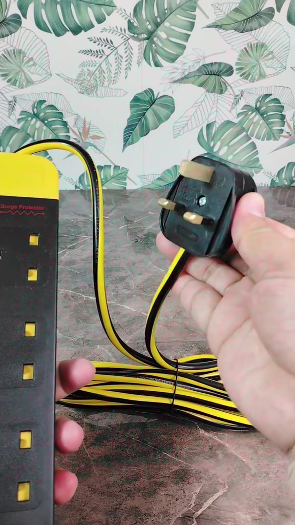 Tangan memegang plug 3 pin Malaysia hitam dengan penutup pin plastik, wayar kuning-hitam berjuntai ke bar extension