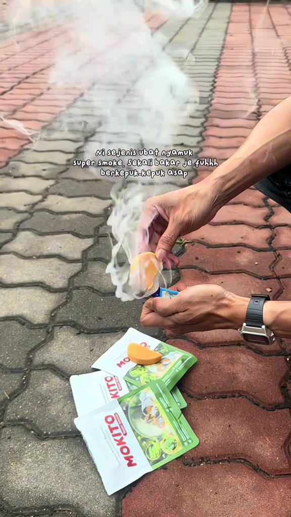 Tangan memegang ubat nyamuk coil yang berasap tebal dengan beberapa paket coil MOKITO di atas lantai berturap