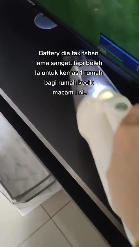 Tangan memegang vacuum handheld cordless putih sedang menyedut habuk atas rak TV hitam