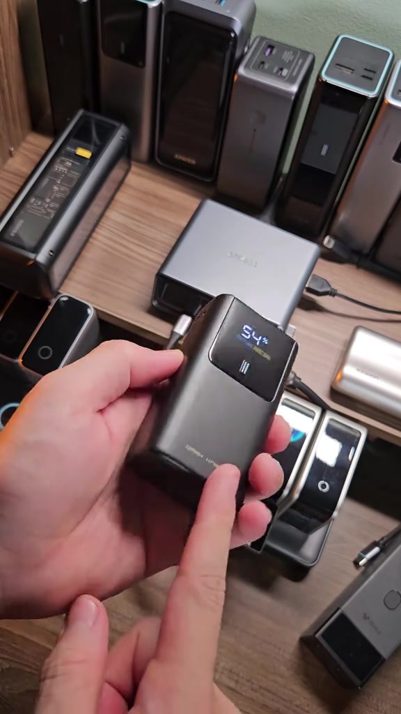 Bàn tay cầm viên CUKTECH 10 mini màn hình hiện 54% pin, phía sau là kệ摆 đầy sạc dự phòng nhiều hãng
