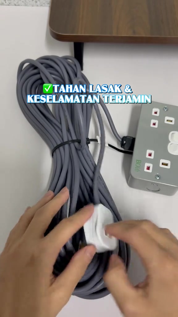 Gulungan wayar kelabu tebal diikat getah di atas meja putih sebelah kotak soket besi, tangan sedang memegang plug putih
