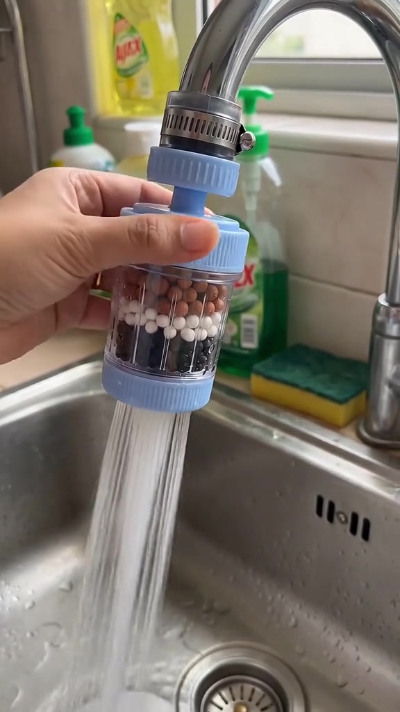 Tangan memegang penapis kepala paip biru berisi lapisan batu tapis dengan air menyembur keluar ke dalam sinki stainless steel