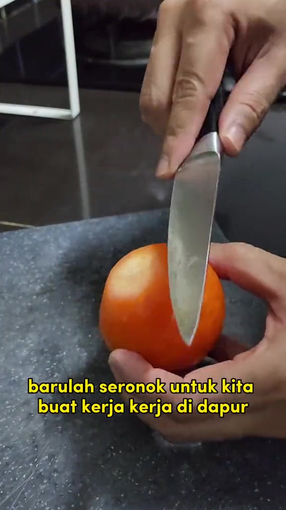 Dua tangan menekan pisau dapur ke atas biji tomato di atas papan potong kelabu