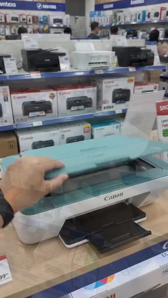 Tangan membuka penutup imbasan printer Canon PIXMA berwarna teal di kaunter kedai, dengan kotak printer di latar belakang