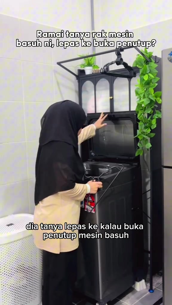 Wanita bertudung dilihat dari belakang membuka penuh penutup mesin basuh top load di bawah modul rak yang tergantung tinggi