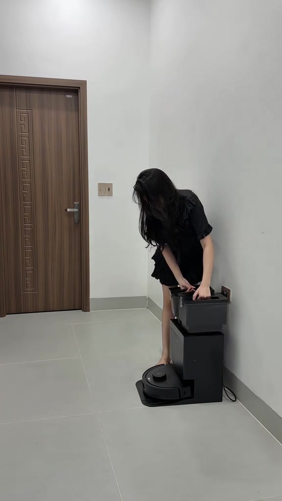 Một người cúi nhấc khay nước khỏi trạm giặt khăn màu đen đặt sát tường, robot đỗ ở chân trạm