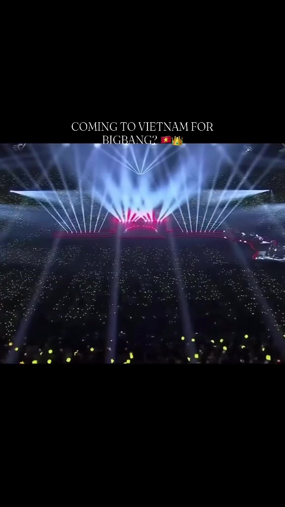 Sân khấu concert BIGBANG nhìn từ xa với chùm tia sáng trắng tỏa lên và biển lightstick vàng lấp lánh trải khắp khán đài